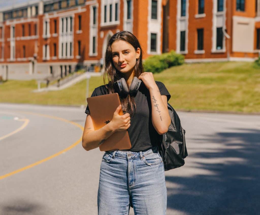 Une jeune étudiante avec des écouteurs se tient avec assurance devant un bâtiment, serrant son ordinateur portable et paraissant inspirée.