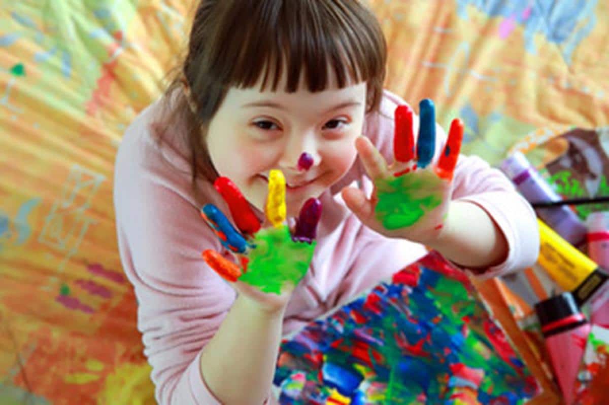 Une jeune enfant trisomique nous sourit en nous montrant ses mains pleines de gouache de couleurs.